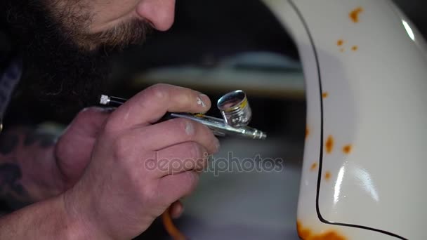 Ο άνθρωπος γένια ενηλίκων στη μαύρη cap ζωγραφική με αερογράφο στο φτερό στο προσαρμοσμένο γκαράζ αυτοκινήτων — Αρχείο Βίντεο