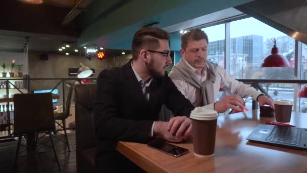Dos compañeros de trabajo hombre adulto se pone gafas y con su joven compañero de trabajo discute un proyecto con un ordenador portátil en un café — Vídeos de Stock