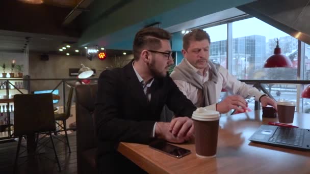 Zwei Mitarbeiter erwachsener Mann setzt Brille auf und diskutiert mit seinem jungen Mitarbeiter ein Projekt mit Laptop in einem Café — Stockvideo