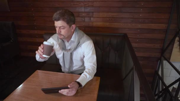 Adult stilig man arbetar på en surfplatta och dricker kaffe på ett kafé — Stockvideo