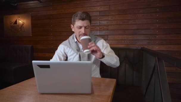 Hombre guapo adulto en la cafetería bebe sonrisas de café cerró el portátil y comienza a bailar mostrando el éxito del trabajo realizado — Vídeos de Stock