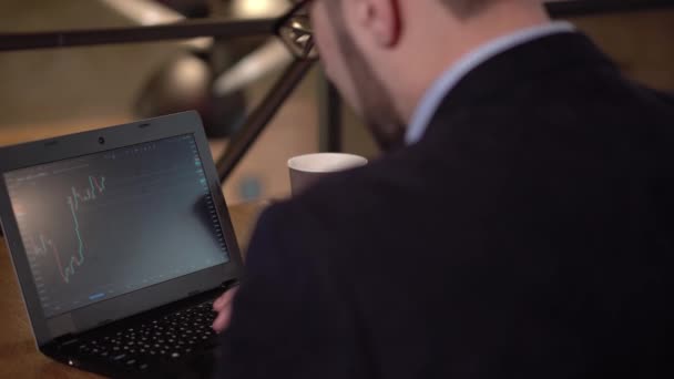 Hombre con gafas estudia gráficos comerciales en un ordenador portátil mientras está sentado en un café — Vídeo de stock