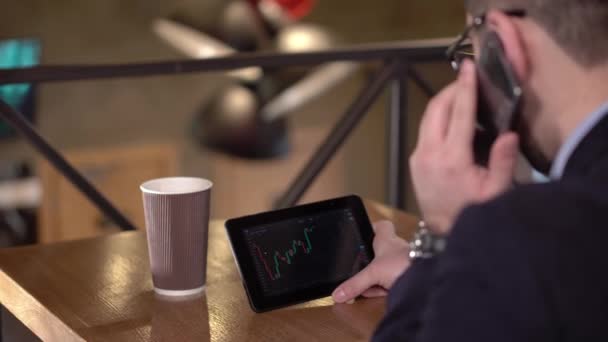 Jovem monitora negociação criptomoeda no tablet e fala telefone em um café — Vídeo de Stock