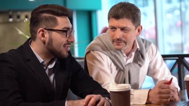 Reifer gutaussehender Mann und ein junger Mann mit Brille unterhalten sich in einem Café — Stockvideo