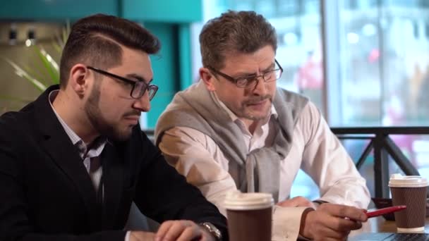 Twee collega's volwassen man zet op bril en met zijn jonge collega bespreekt een project met een laptop in een café — Stockvideo