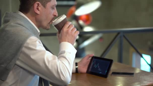 Volwassen knappe man drinkt koffie en controleert cryptocurrency handel op tablet in een café — Stockvideo