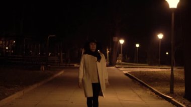 Şapkalı genç esmer kız gece parkta yürüyor ve telefonunu kontrol etmek başlar
