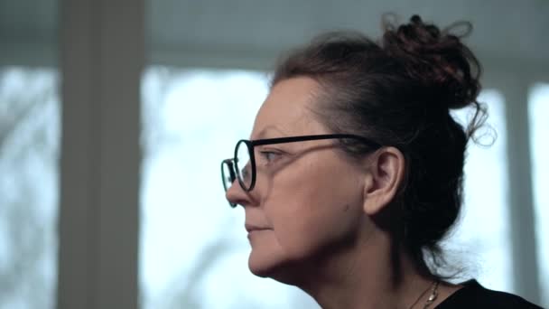 Mujer morena madura en gafas mira hacia adelante en frente de la ventana interior — Vídeos de Stock