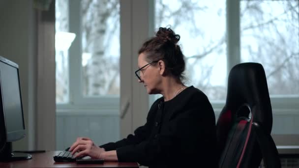 Зрелая брюнетка в очках работает за компьютером в офисе сидя в кресле — стоковое видео