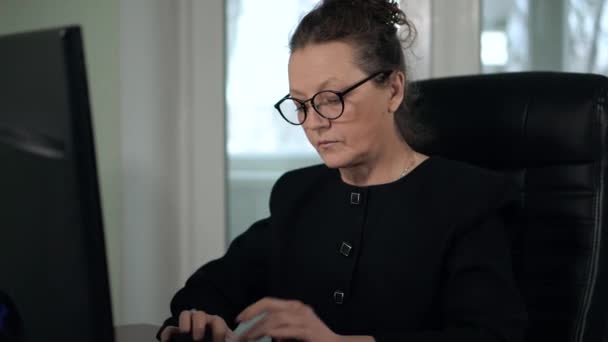 Mature woman brunette in glasses and black suit typing on computer in office sitting in front of window — Stock Video