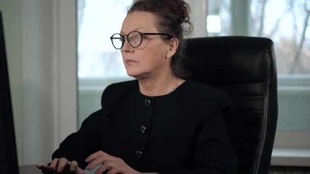 Зріла жінка брюнетка в окулярах і чорний костюм друкує на комп'ютері і приймає телефонний дзвінок — стокове відео