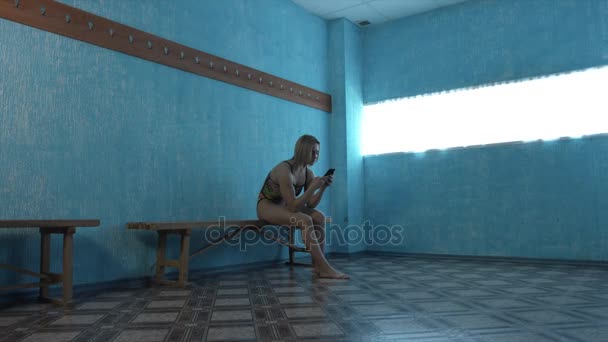 Девушка в купальнике сидит в раздевалке и смотрит на смартфон — стоковое видео
