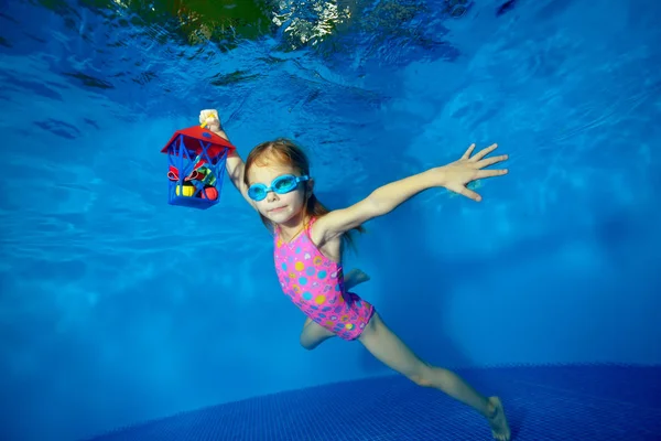 Pequeño niño feliz nada bajo el agua en la piscina con juguete-regalo en las manos sobre fondo azul y mira a la cámara. Retrato. Disparando bajo el agua. Orientación al paisaje — Foto de Stock