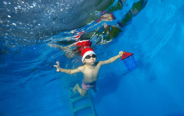 Niño feliz en un sombrero Santa Claus con un regalo en la mano nada bajo el agua en la piscina con los brazos extendidos sobre un fondo azul — Foto de Stock