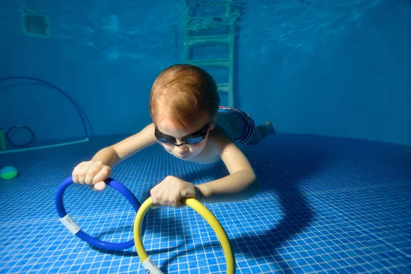 El niño nada y entrena bajo el agua en la piscina y recoge juguetes del fondo de la piscina. Disparos bajo el agua — Foto de Stock