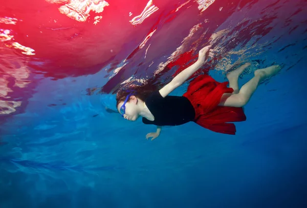 Feliz niña nadando y jugando bajo el agua sobre un fondo azul en una falda roja . —  Fotos de Stock