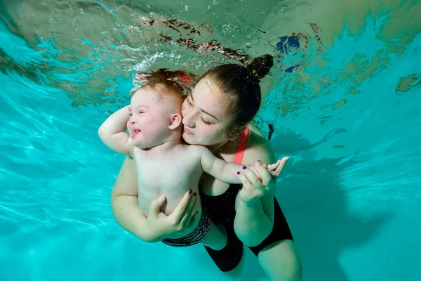 Un bebé en un pañal junto con una madre nada bajo el agua en la piscina, acurrucándose y mirando hacia un lado . — Foto de Stock