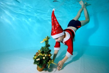Happy little boy in a cap Santa Claus dives to the bottom of the pool to get a Christmas tree clipart