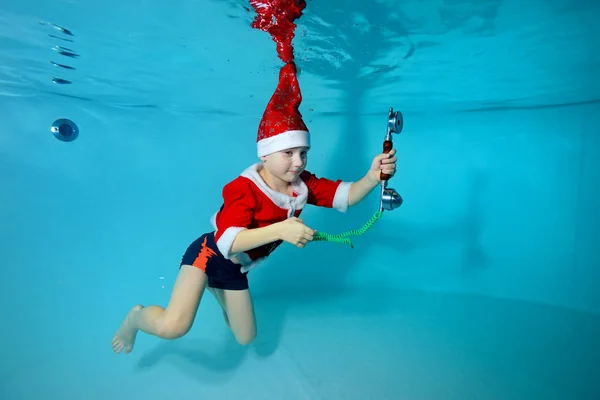 Jongetje in Kerstman hoed drijft onder water in het zwembad en met de telefoon in zijn hand. Portret. Fotograferen onder water. Horizontale oriëntatie — Stockfoto