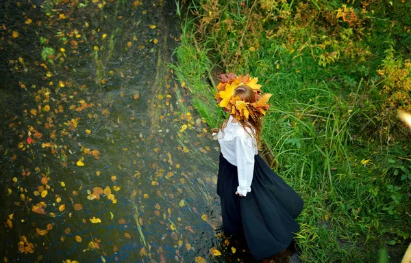 Beautiful young girl wearing a wreath of autumn leaves in a white shirt and black skirt enters the river. The view from the top. Horizontal orientation