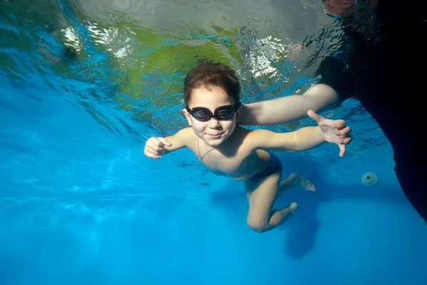 Sports little boy swims with a trainer underwater in the pool on a blue background, looking at camera and smiling. Portrait. Shooting underwater. Landscape orientation