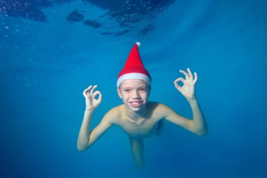 Çocuk havuzunda sualtı Noel Baba'nın kırmızı şapkalı, gülümsüyor, yüzüyor ve kameraya doğru görünüyor. Portre. Su altında çekim. Yatay Yönlendirme