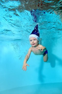 Santa Claus şapka mavi gözlü bebek su altında mavi bir arka plan üzerinde bir havuzda yüzüyor. Portre. Su altında çekim. Dikey yönlendirme
