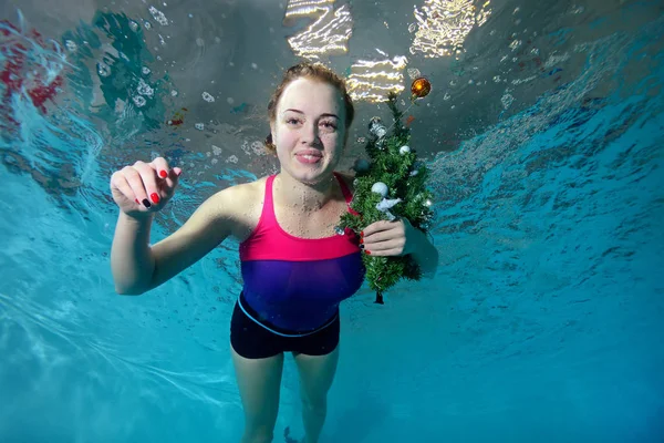 Cute young girl swims underwater in the pool on a blue background with a Christmas tree with a toy in hand, looking at camera and smiling. Portrait. Horizontal orientation. A view from under the water