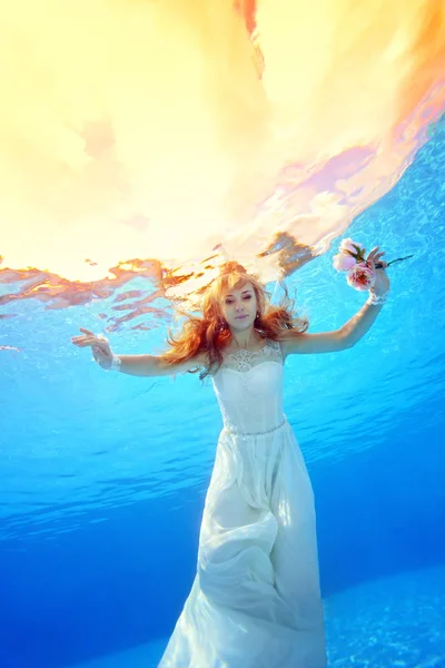 Belle fille en robe de mariée nage sous l'eau dans la piscine au coucher du soleil avec des fleurs dans ses mains. Portrait. Vue verticale. Tir sous l'eau au fond — Photo