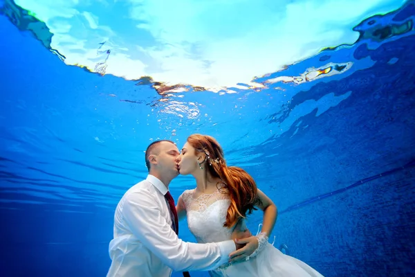 La novia y el novio en vestidos de novia abrazan y besan bajo el agua en la piscina sobre un fondo azul. Primer plano. Orientación al paisaje. Disparos bajo el agua en la piscina — Foto de Stock