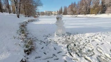 Kardan adam bir şehirde Park nehir üzerinde bir güneşli kış gününde duruyor. 4k, 25fps