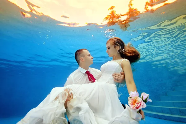 Amour sous-marin un homme tenant sa mariée dans une robe de mariée sur le fond d'un coucher de soleil tropical et la regarde — Photo