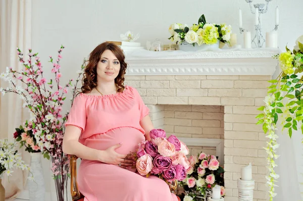 Chica embarazada en vestido rosa sentada en una silla en el estudio, sosteniendo las flores en su mano y mirando a la cámara — Foto de Stock