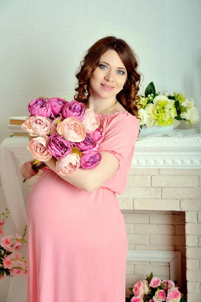 Hermosa joven embarazada en un vestido rosa de pie en el estudio con flores y mirando a la cámara —  Fotos de Stock