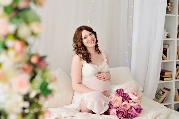 Una joven embarazada en lencería blanca sentada en la cama, pone las manos en el vientre mirando a la cámara y se ríe — Foto de Stock