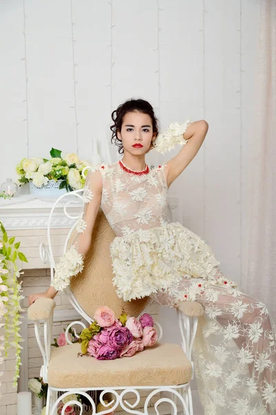 Gyönyörű ázsiai lány fehérnemű jelentő Studio egy vintage szék, egy csokor virágot, és nézi a kamerát — Stock Fotó