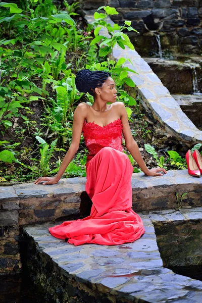 African American girl in a red dress, with dreadlocks, sitting on the stones in the Park on a background of green plants and looking to the side.