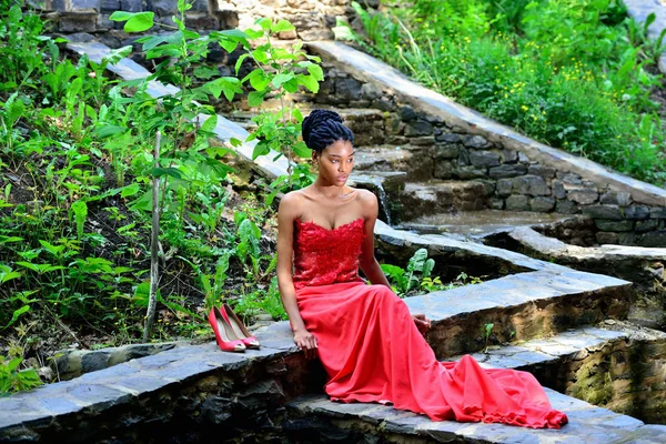 Femme afro-américaine assise dans le parc posant sur fond de plantes vertes sur les rochers dans une robe rouge avec dreadlocks — Photo