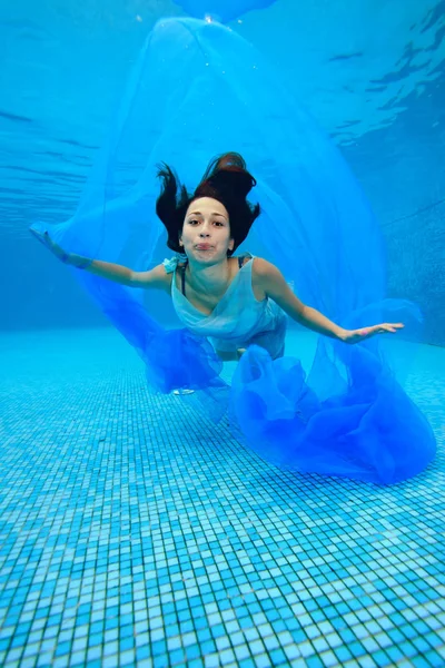 Het tienermeisje in de jurk onderwater duiken op de bodem van het zwembad, speelt met een blauw doek en kijkt naar de camera. Portret. Fotograferen onder water — Stockfoto
