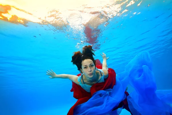 Una adolescente con un paño rojo y azul se sumerge bajo el agua en el fondo de la piscina contra el fondo de luces brillantes y mira a la cámara. Retrato. Disparos bajo el agua —  Fotos de Stock