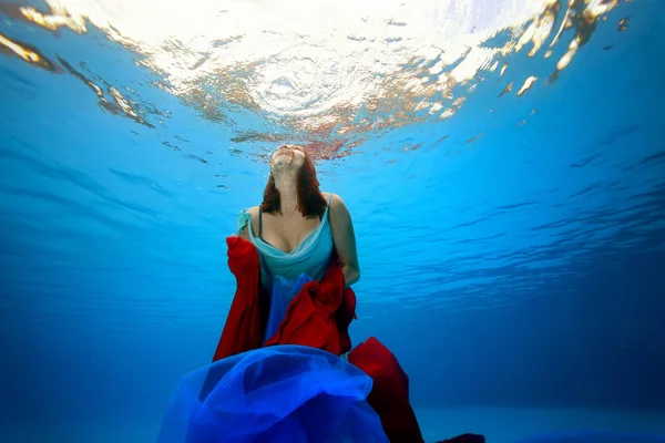 Dziewczyna z czerwonej i niebieskiej tkaniny w jej rękach pływa pod wodą na powierzchni morza od dołu. Portret. Fotografowanie pod wodą — Zdjęcie stockowe