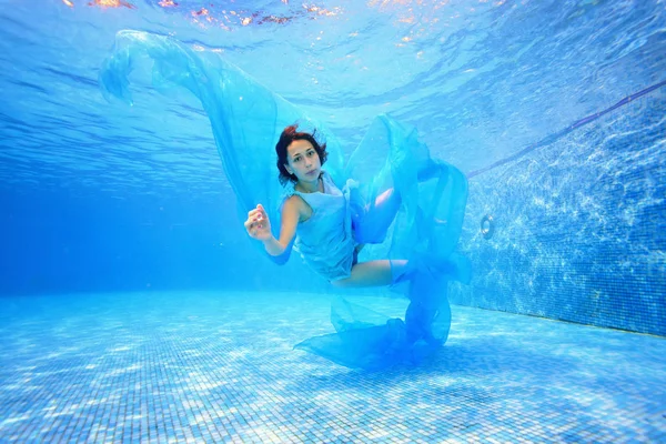 Een tienermeisje in een blauwe jurk en met een blauw doek in haar hand onder water zwemt in het zwembad tegen een blauwe achtergrond en kijkt naar de camera. Portret. Fotograferen onder water — Stockfoto