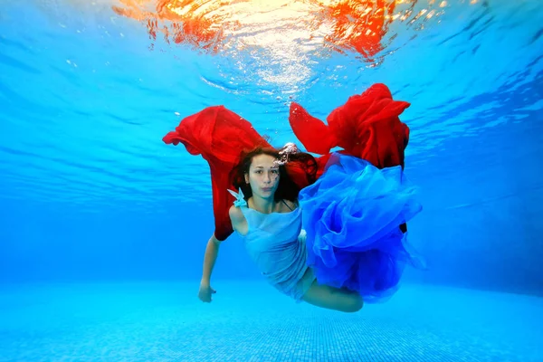 Piękna dziewczyna w sukience pływa pod wodą w basenie, odgrywa szmatką czerwony i niebieski i patrzy w kamerę. Fotografowanie pod wodą. — Zdjęcie stockowe