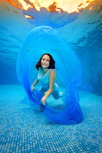 Flickan i klänning poserar under vattnet på botten av poolen, leker med en blå duk, kameran och tittar och ler mot bakgrund av starkt ljus. — Stockfoto