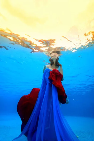 Het meisje met een rode en blauwe doek in haar handen ijslollie opwaarts uit de bodem van het zwembad naar de oppervlakte. Fotograferen onder water. — Stockfoto