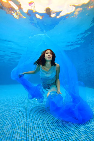 Het meisje in de jurk zwemt en vormt onder water op de bodem van het zwembad, speelt met een blauw doek en kijkt naar de camera tegen de achtergrond van fel licht. Portret. Fotograferen onder water — Stockfoto