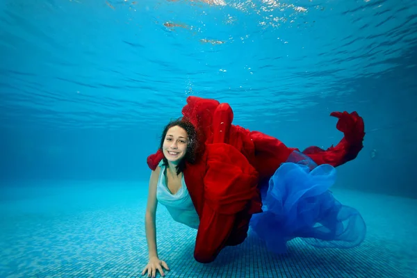 穿着裙子的少女在泳池的底部游泳 玩着红蓝布 看着摄影机大笑 在水下射击 景观定位 — 图库照片