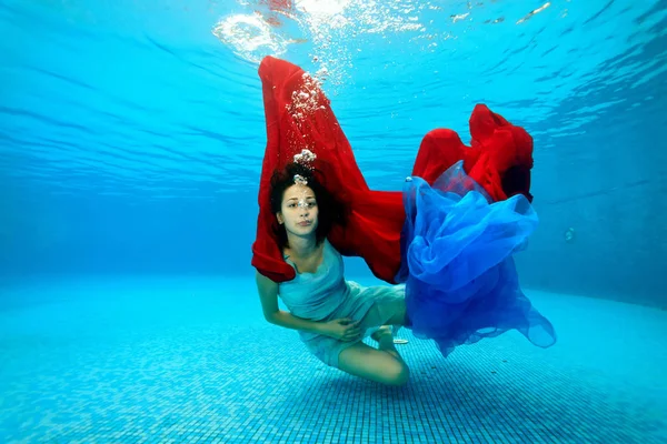 La chica juega bajo el agua en el fondo de la piscina con un paño rojo y azul y mira a la cámara. Retrato. Disparos bajo el agua . — Foto de Stock