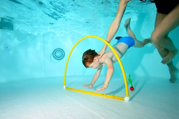 The coach helps the little boy swim underwater at the bottom of the pool through the Hoop. Shooting underwater from the bottom. Horizontal view