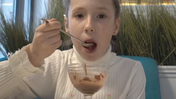 En liten flicka i en vit klänning och med fräknar på hennes ansikte sitter på ett café och äter glass från ett glas Cup. Underifrån. — Stockvideo